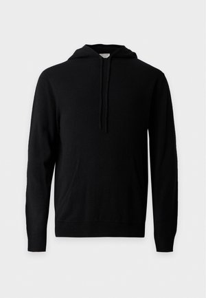 Homme portant un sweat à capuche noir, un pantalon gris foncé et des chaussures noires, debout devant un fond blanc uni avec les mains détendues.