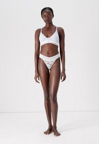 Svetlo sivo bralette z nastavljivimi naramnicami in sprednjim zapiranjem, v kombinaciji z belim čipkastim tangicami, ki imajo valovite robove.