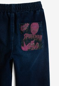 Tumed sinised denim püksid musta ja roosa Spider-Mani graafika plaastriga tagakotil, millel on koomiksilaadsed elemendid ja aksendid.