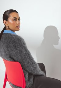 Suéter gris de felpa hecho de material texturizado, llevado sobre una camisa de cuello azul; sentado en una silla roja brillante frente a un fondo claro.