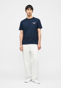 Άνδρας που στέκεται φορώντας μπλε ναυτικό t-shirt Polo Sport, λευκά παντελόνια με φαρδιά πόδια και λευκά αθλητικά παπούτσια, σε απλό φόντο.