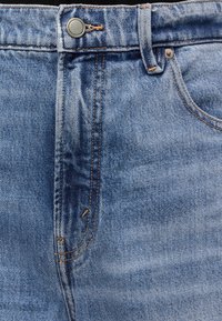 Κοντινή λήψη της μέσης από μπλε τζιν παντελόνι με μεταλλικό κουμπί, θηλιά ζώνης, πορτοκαλί ραφές και λεπτομέρεια μπροστινής τσέπης.