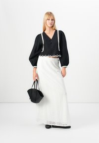 Blouse noire raccourcie avec des boutons et des détails en broderie blanche, associée à une longue jupe blanche avec des motifs texturés. Le mannequin porte un sac noir.