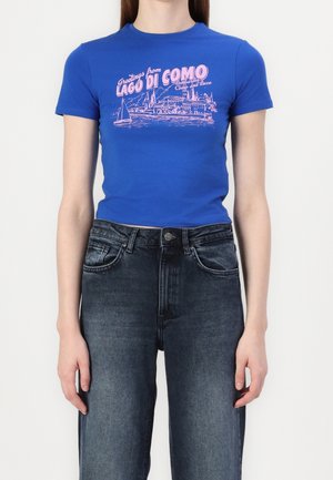 Crop top bleu ajusté avec texte rose "Greetings from Lago di Como" et motif scénique, porté avec un jean noir délavé taille haute.