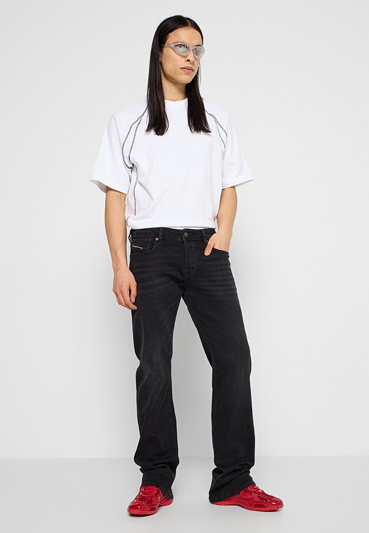 Person mit langen schwarzen Haaren, die klare Brillen trägt, ein weißes kurzärmeliges Hemd, schwarze Jeans und rote Sneakers an einer weißen Wand steht.