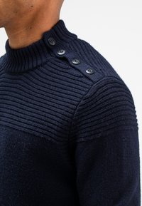 Navyblauw gebreid sweater met een hoge ribgebreide kraag, textuurstrepen op de schouders en vier zwarte knopen op de linker schouder.