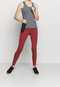 Top sportivo grigio testurizzato, leggings bordeaux con striscia laterale, sneakers nere, mentre tiene una bottiglia d'acqua nera. Abbigliamento sportivo semplice in un ambiente neutro.