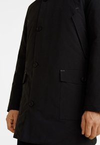 Hombre vestido con un abrigo negro abotonado con bolsillos de solapa, de pie con las manos relajadas a los lados sobre un fondo blanco.