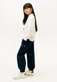 Felpa bianca con scollatura a volant, pantaloni cargo blu navy con tasche e sneakers bianche con accenti leopardati. Capelli neri lunghi.