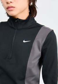 Top sport negru cu fermoar, cu guler înalt, panouri laterale gri și logo alb Nike. Fabricat dintr-un material neted și elastic, potrivit pentru îmbrăcăminte activă.