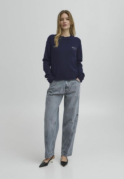 Marineblå strikket genser med rund hals og lite logo, kombinert med lys grå baggy jeans og svarte spisse hæler.
