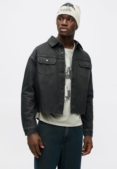 Črna skrajšana denim jakna z dvema žepoma na prsih, z gumbnim zapiranjem in kontrastnim šivom, oblečena čez svetlo grafično majico in temne kavbojke.