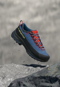 Blå tursko med svart såle, røde detaljer og grå snørebånd. Har en teksturert overdel og lett design for utendørsaktiviteter.