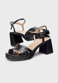 Sandales à talons noirs avec un bout ouvert courbé, dotées de larges brides et d'une boucle à la cheville. Talon épais en bloc et texture en cuir lisse et brillant.