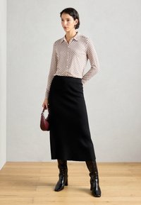 Kvinna som bär en mönstrad långärmad blus, svart midi kjol, svarta klackboots, håller en liten vinröd handväska, står på trägolv.