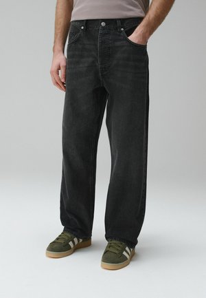 Luźne czarne jeansy męskie noszone z zielonymi zamszowymi sneakersami z białymi paskami, stojący z jedną ręką w kieszeni.