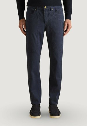 Uomo che indossa pantaloni casual blu scuro con ampie tasche frontali, bottone dorato, scarpe nere con suole beige e maglia nera a maniche lunghe.