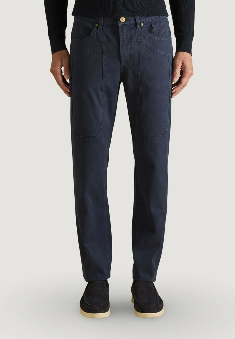 Hombre vistiendo pantalones casuales azul marino oscuro con grandes bolsillos delanteros, botón dorado, zapatos negros con suelas beige y camiseta negra de manga larga.