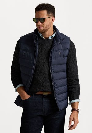 Gilet matelassé bleu marine superposé à un pull à col roulé noir, associé à une chemise à col bleu clair, un jean foncé et des lunettes de soleil.