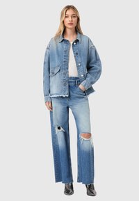 Ξανθιά γυναίκα που φοράει ανοιχτό μπλε φαρδιά τζιν μπουφάν και τζιν με σκισίματα και πατς, μαζί με μαύρα μυτερά μποτάκια, όρθια μπροστά σε απλό φόντο.