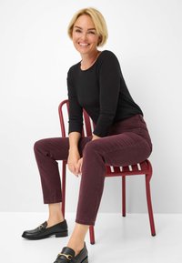 Schwarzes Langarmshirt, burgunderfarbene, schmal geschnittene Hose und schwarze Slipper mit goldener Hardware, auf einem roten Stuhl vor einem weißen Hintergrund sitzend.