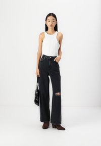 Svarta högmidjade vida jeans med ett slitage vid knäet, tillsammans med en figurnära vit tanktopp och bruna spetsiga ankelskor med klack. Svart väska.