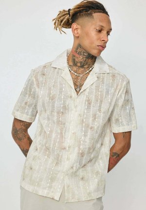 Homme aux dreadlocks attachés en arrière, portant une chemise beige semi-transparente à manches courtes et un collier en argent, exhibant des tatouages sur le cou et les bras.