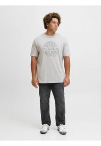 Camiseta gris claro con un gráfico circular impreso en gris oscuro, combinada con jeans oscuros y zapatillas blancas con acentos en gris y negro.