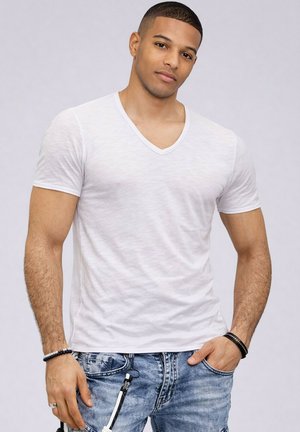 Jeune homme aux cheveux courts portant un T-shirt blanc à col en V, un jean bleu délavé, et des bracelets noirs et blancs aux deux poignets.
