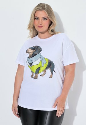 Donna bionda con una t-shirt bianca con un bassotto da cartone animato con occhiali da sole, sciarpa e giacca gialla, abbinata a pantaloni in pelle neri.