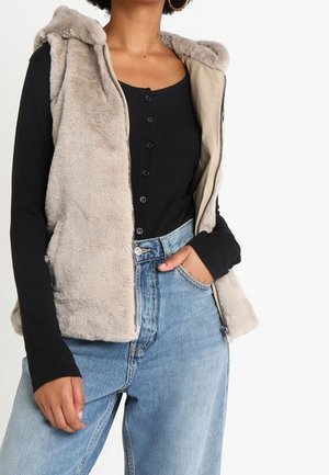 Femme portant un gilet sans manches beige en fausse fourrure zippé sur un haut noir à manches longues boutonné et un jean bleu clair taille haute, une main dans la poche.