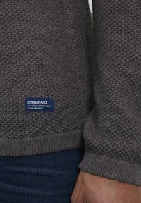 Grijze gebreide trui met een textuurpatroon, geribbelde manchetten en onderzoom. Heeft een blauw label met logo en tekst nabij de onderzoom.
