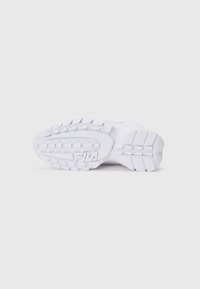Semelle d'une chaussure de sport blanche avec des profondeurs profondes et le logo FILA embossé près de la zone de la chaussure, photographiée sur un fond uni.