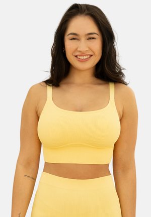 Woman with long dark hair smiling, wearing a yellow ribbed crop top and matching high-waisted bottoms on a plain background.