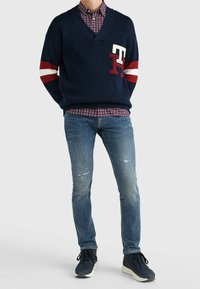 Marinblå tröja med röda och vita randiga ärmar, v-ringad design, prydd med en stor "TH"-logotyp. Kombinerad med mönstrad skjorta och slitna blå jeans.