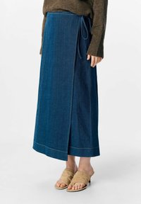 En denim indpakningsskørt i mørkeblå med en sløjfe i taljen, der har kontraststing og en lige silhuet. Parret med lysebrune slidssandaler.