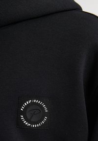 Tissu de sweat à capuche noir avec une surface texturée, présentant un patch en forme de carré avec un texte circulaire blanc et un "P" stylisé au centre.