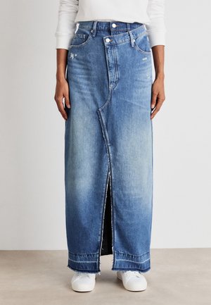 Kvinde iført en lang blå denim nederdel med en asymmetrisk frontslids og hvide sneakers, stående mod en ensfarvet baggrund.