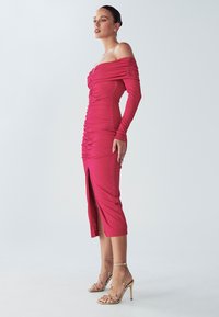 Priliehavé šaty bez ramienok v žiarivej ružovej farbe, s nariasením na trupe a bočným rozparkom. Dopĺňajú ich zlaté sandále na vysokom opätku s remienkami.