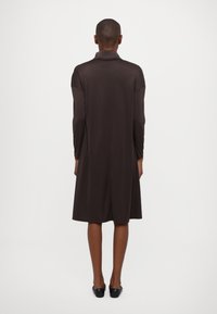 Abito lungo di colore marrone scuro realizzato in tessuto morbido, con maniche lunghe e una vestibilità ampia, dotato di colletto e che arriva a metà polpaccio.