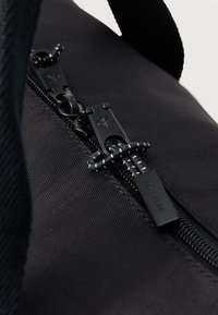 Zwarte nylon tas heeft een gladde textuur met een merkenrits en contrasterende stiksels langs de ritssluiting.