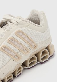 Scarpa da ginnastica beige con corpo in mesh, suola in gomma con cilindri cavi e tre strisce di plastica sovrapposte con borchie.