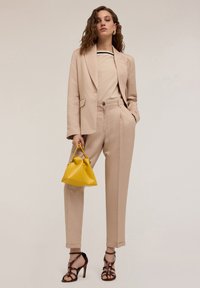 Beżowa marynarka wykonana na miarę z dopasowanymi spodniami, z podkreśloną talią i bocznymi kieszeniami. Żółta torebka i sandały na wysokim obcasie dopełniają całość.