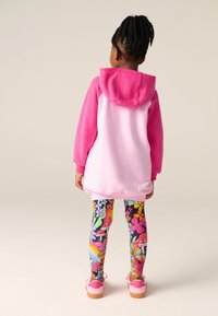 Rózsaszín kapucnis pulóver puha textúrával, kenguruzsebbel és kapucnival. Színes, mintás leggingsszel és rózsaszín cipővel párosítva.