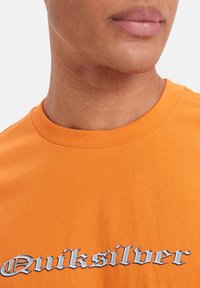 Oranžové bavlněné tričko s kulatým výstřihem, s kontrastním stříbrným logem "Quiksilver" v tučném, stylizovaném písmu.
