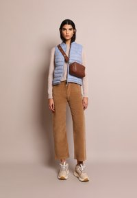 Gilet trapuntato azzurro chiaro sopra un maglione beige, abbinato a pantaloni corduroy beige a gamba larga e sneakers beige. Borsa a tracolla marrone con cinturino.