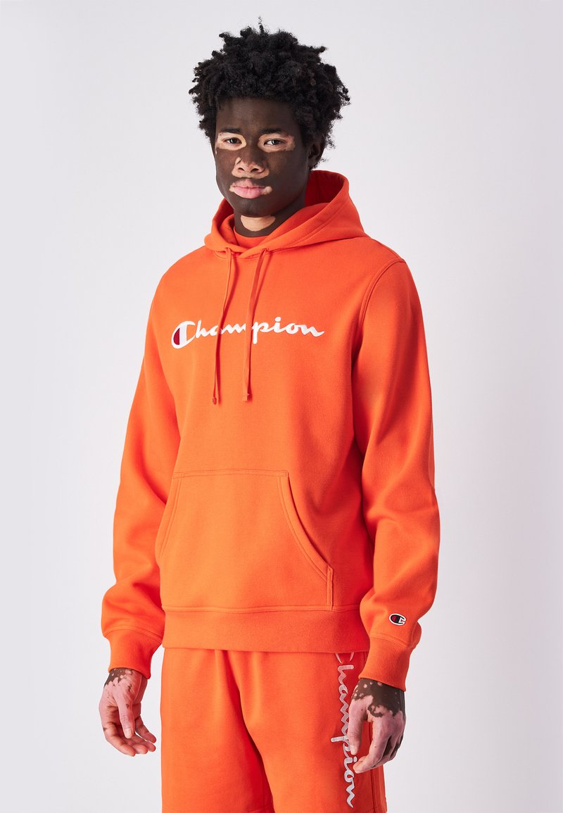 Sudadera con capucha de un brillante color naranja, con un bolsillo frontal y el logo en blanco de "Champion". Puños y bajo acanalados, fabricada en material suave.