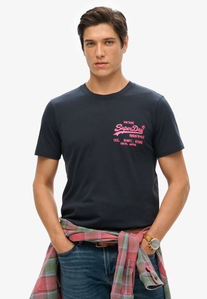 Granatowa bawełniana koszulka z krótkim rękawem, z różowym tekstem graficznym na piersi. Koszula w kratę owinięta w talii dodaje kolorowego kontrastu.