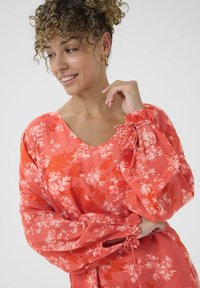 Bluse mit Blumenmuster in Koralle, mit V-Ausschnitt, langen Puffärmeln und gerafften Manschetten. Sie besteht aus einem leichten, strukturierten Stoff mit pinken und roten Blumenmustern.
