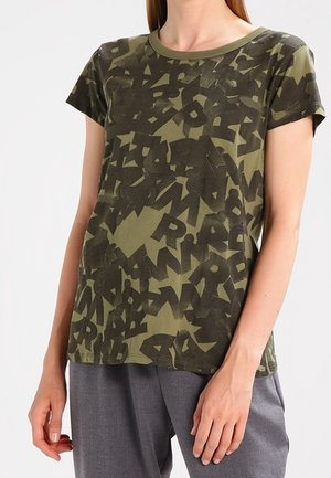 Woman wearing an olive green T-shirt with black abstract letter patterns and gray pants, standing with hands relaxed by sides.